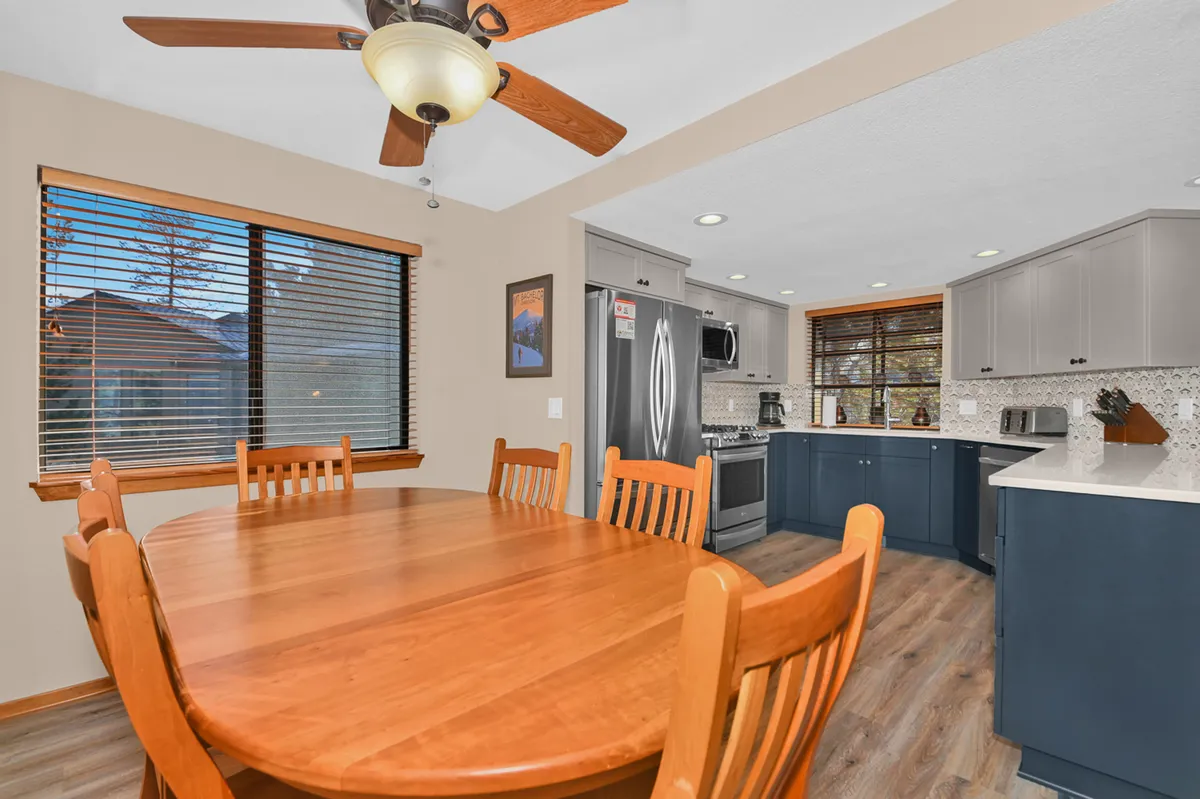 The image captures a cozy dining area adjacent to a modern kitchen, with a substantial dining table set for group meals. Large windows provide a glimpse of the outdoors and flood the space with natural light, creating a sociable, inviting atmosphere ideal for gatherings with friends or couples.