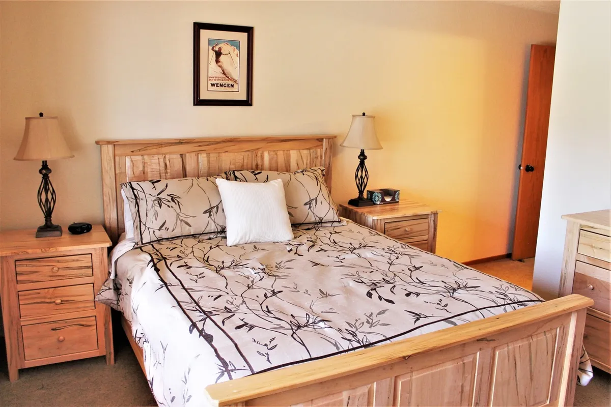 A serene bedroom featuring a wooden bed frame with a decorative floral bedspread, flanked by nightstands with lamps. Soft natural light filters into the room, creating a peaceful atmosphere ideal for relaxation and restful sleep.