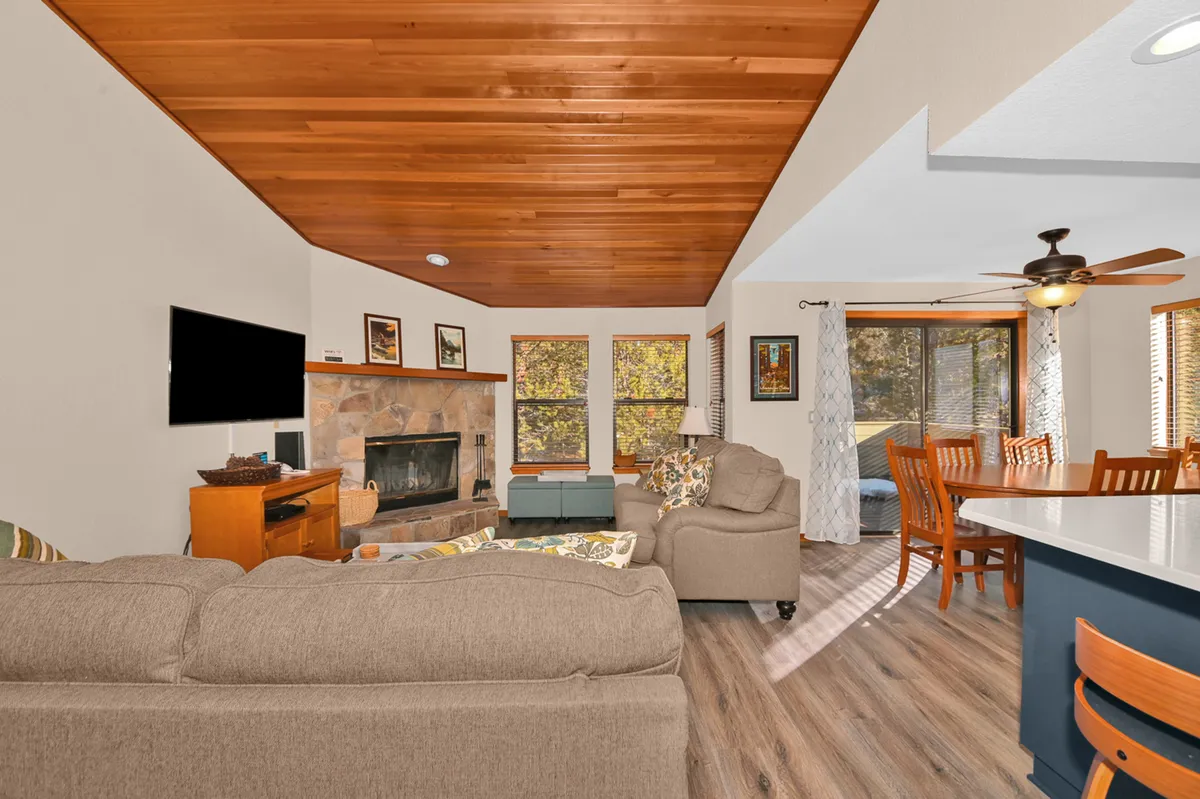 A sunlit open-concept living area featuring a cozy fireplace, plush sectional sofa, inviting dining space, and expansive windows with woodland views. The warm, stylish furnishings and natural light create an inviting setting for relaxation and family gatherings.