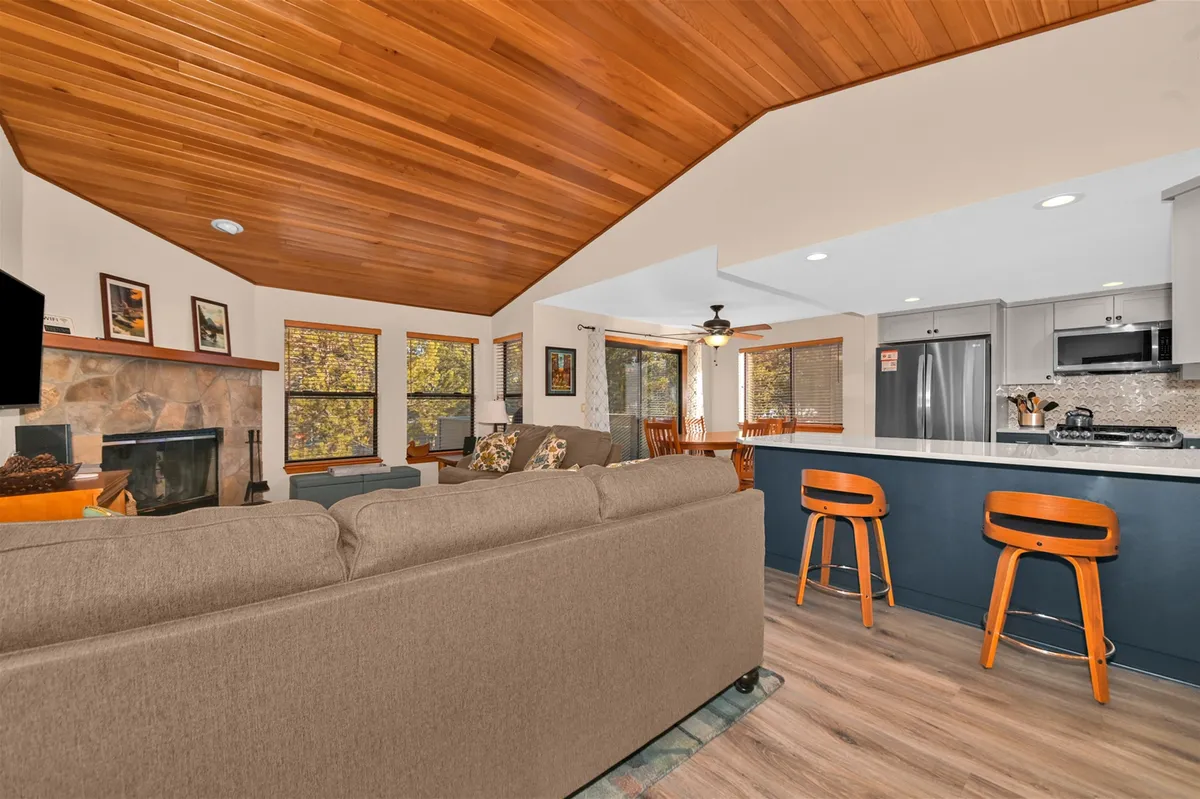 A welcoming living space featuring a striking stone fireplace, warm wooden ceiling, and a kitchen island in view. The area is arranged for relaxation and entertaining, with comfortable furnishings that invite guests to unwind together after a day of activities.