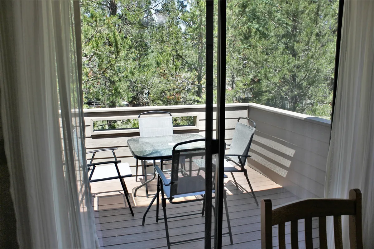 A serene outdoor dining area on a deck, featuring a glass table with metal chairs surrounded by green trees and a wooden railing. The scene captures the peaceful ambiance of the complex’s outdoor spaces, suggesting summer gatherings and relaxation in nature.
