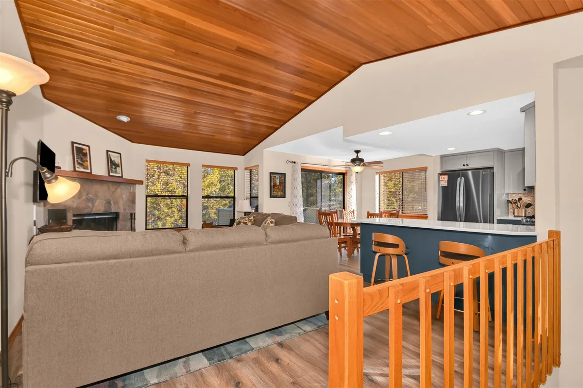 This inviting open-concept space seamlessly connects the living room, kitchen island, dining area, and a prominent wood-burning fireplace. Sunlight streams through large windows, creating a warm ambiance ideal for gatherings, relaxation, and entertaining. Plush seating and tasteful decor invite guests to unwind, while the fireplace anchors the heart of the home.